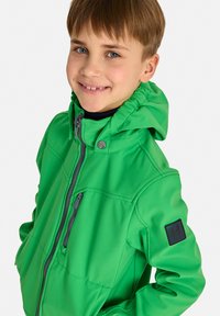 Giacca softshell verde con cappuccio, caratterizzata da dettagli grigi delle zip e un patch con logo sulla manica sinistra; texture liscia e taglio aderente.