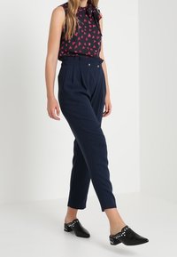 Marineblauwe hoog taille broek met plooien, gecombineerd met een zwarte mouwloze blouse met rode hartpatronen, en zwarte muiltjes met studs.