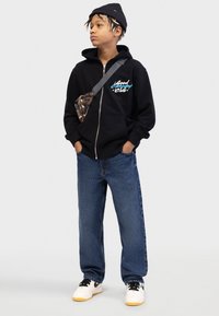 Μαύρο φούτερ με φερμουάρ και λογότυπο, με μπλε λεπτομέρειες, σε συνδυασμό με μπλε τζιν και λευκά αθλητικά παπούτσια με κίτρινες σόλες. Μαύρος πλεκτός σκούφος με υφή.