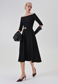 Schwarzes Off-Shoulder-Kleid mit Dreiviertelärmel, tailliert mit einem goldenen Gürtel, kombiniert mit langen schwarzen Handschuhen und burgunderfarbenen Slingback-Absätzen.