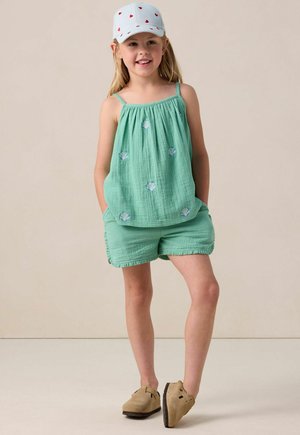 Débardeur en coton vert avec broderie en forme de coquillage, shorts assortis, et une casquette bleu clair avec des motifs de cœurs rouges. Des chaussures beiges complètent la tenue.