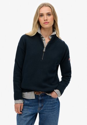Navy pullover sweater with a zip neckline, ribbed cuffs, and contrasting striped shirt underneath, paired with blue denim jeans.