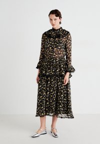Kvinne som har på seg en svart gjennomsiktig blomstret langermet bluse og matchende midikjole med volanger, kombinert med sølvfargede Mary Jane-flate sko, står mot en hvit vegg.