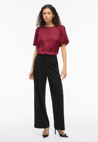 Blouse en satin bordeaux à manches bouffantes, associée à un pantalon large noir et des chaussures noires à talons hauts avec un accent de chaîne dorée.