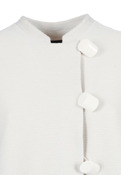 Wit textiel kledingstuk met een opstaande kraag en drie grote, glanzende, niet-centrale witte knopen langs de voorzijde van de sluiting.