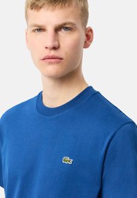 Camiseta de cuello redondo azul hecha de algodón, con un pequeño logo de cocodrilo verde en el lado izquierdo del pecho, textura suave y corte estándar.