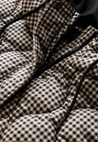 Gestepptes Innenfutter einer Daunenjacke in beigem und marineblauem Karo-Muster, mit einem schwarzen Reißverschluss, Druckknöpfen und glatter Textur.