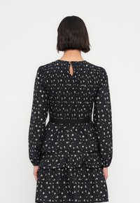 Robe noire à fleurs avec manches longues, corsage smocké, ouverture en trou de serrure dans le dos, et ourlet à volants à étages, ornée de petits motifs blancs et bleus.