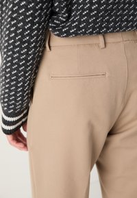 Pantalones beige ajustados con una textura suave y un bolsillo trasero. Combinados con un suéter oscuro de patrón con acentos blancos.