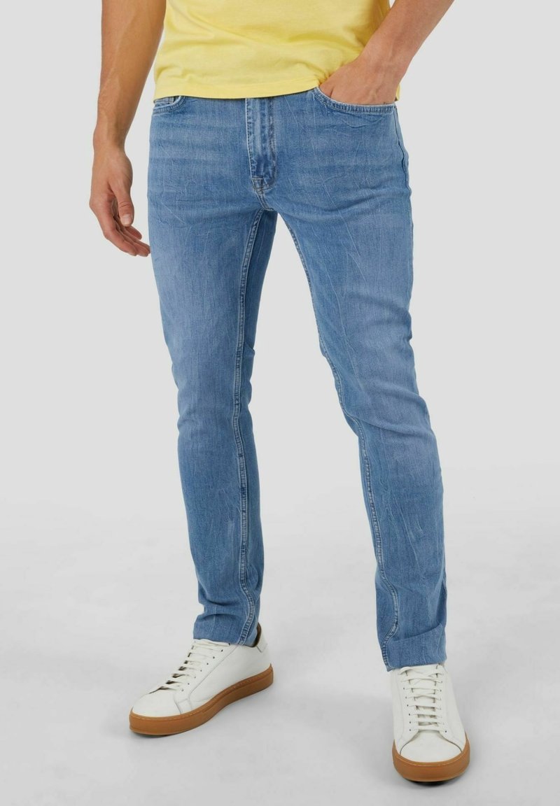 Homme portant un jean slim bleu clair, des baskets blanches avec des semelles marron et une chemise jaune, debout avec une main dans la poche.