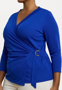 Blue wrap top with long sleeves, featuring gathered fabric detail and gold circular accent at the waist. Smooth, stretchy material.