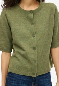 Oliwkowozielony kardigan z krótkim rękawem i okrągłym dekoltem, zapinany na sześć guzików, wykonany z miękkiego, teksturowanego dzianego materiału. Noszony na niebieskich jeansach z denimu.