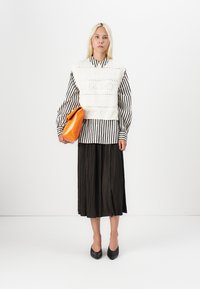 White knitted vest over a black and white striped shirt, paired with a pleated black skirt and an orange bag. Black heeled shoes complete the look.