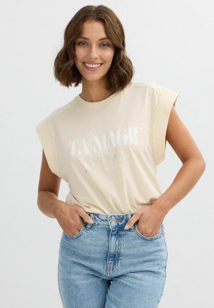 Camiseta sin mangas de un beige claro con texto metálico "TA MAGIE Paris," combinada con jeans de tiro alto azul claro. Tejido suave, ajuste relajado.