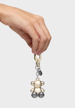 Mano sosteniendo un llavero de oso de peluche de metal dorado y plateado con un pequeño colgante circular unido por una cadena de eslabones, sobre un fondo liso.