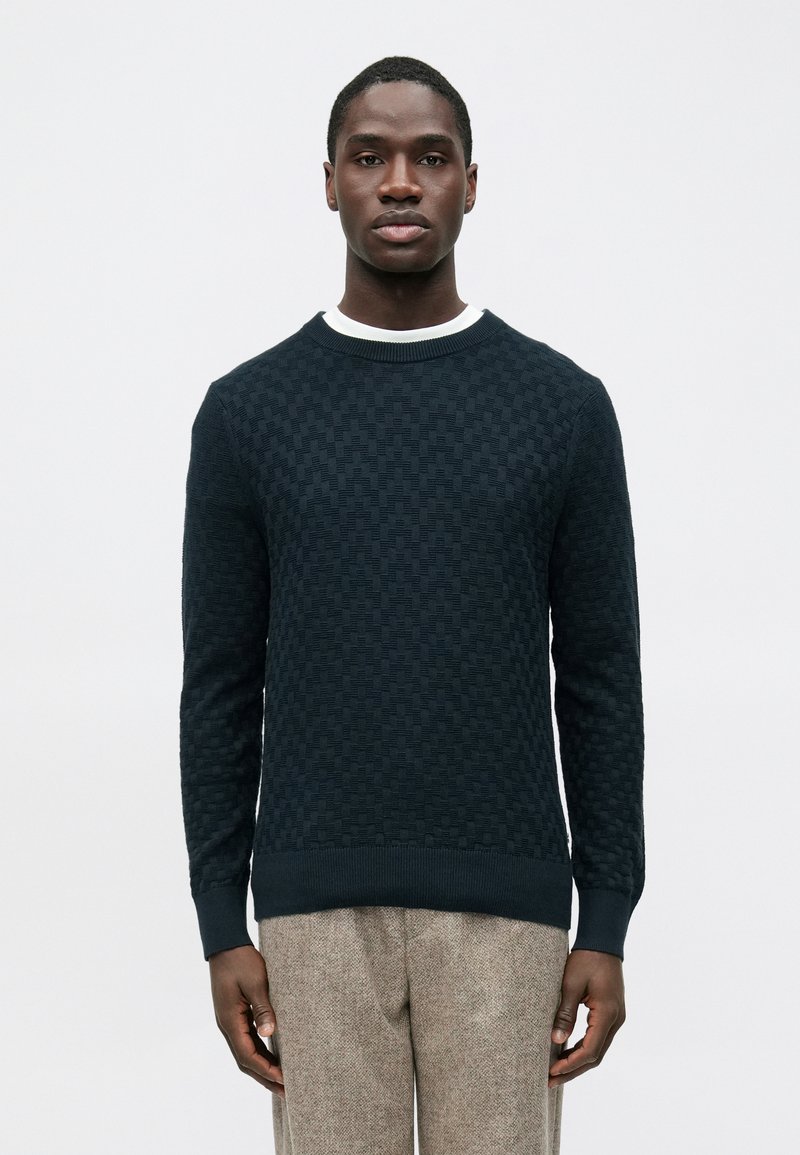 Pull bleu foncé texturé avec un motif à carreaux, col et poignets côtelés, associé à un pantalon texturé marron clair.