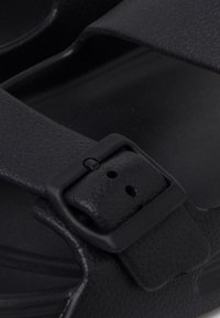 Sandale noire texturée avec une large bride et une boucle carrée noire sur le pied.