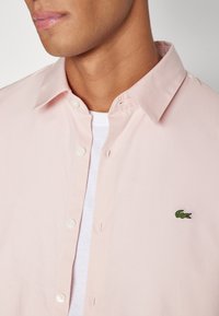 Cămașă din bumbac roz deschis, cu guler tip button-down, două butoane vizibile și un logo brodat cu un crocodil verde pe piept.