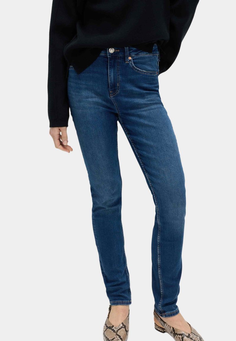 Persona con jeans ajustados azul oscuro, top de manga larga negro y zapatos planos con estampado de serpiente, de pie frente a un fondo claro.