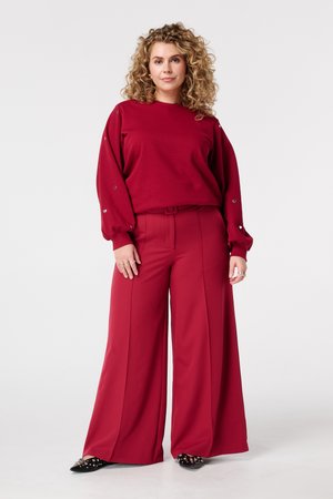 Vrouw met krullend haar die een rood langarmig shirt met hartvormige details draagt en wijde rode broek, staand tegen een effen achtergrond.