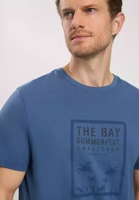 Niebieska bawełniana koszulka z krótkim rękawem, z ciemnym, nadrukowanym prostokątnym wzorem z tropikalnymi palmami oraz napisem "Wyzwanie Letniego Festiwalu Zatoki".