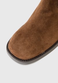 Bruine suede enkellaars met een ronde teen, voorzien van een zwarte gestikte rand en een gladde zool, die een zachte textuur en eenvoudig ontwerp laat zien.