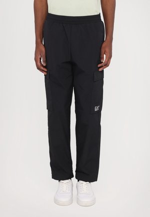 Homme portant un pantalon cargo noir avec des poches latérales et des baskets blanches, debout devant un fond uni.