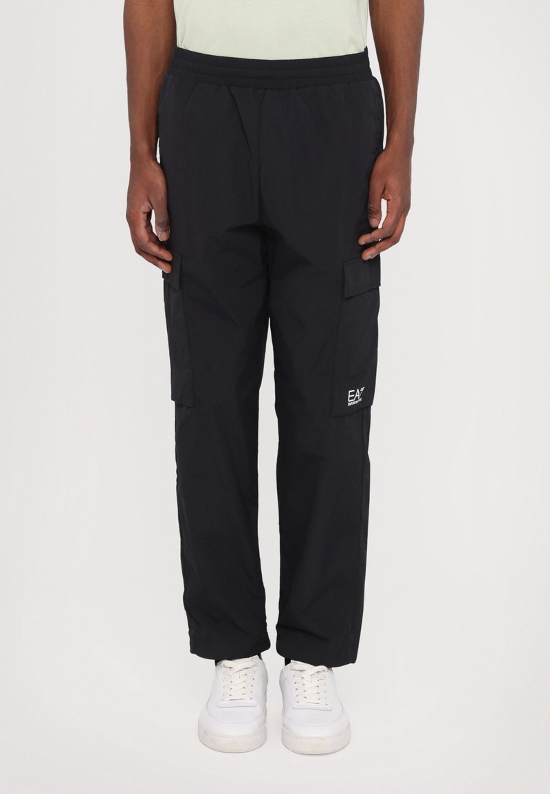 Homme portant un pantalon cargo noir avec des poches latérales et des baskets blanches, debout devant un fond uni.