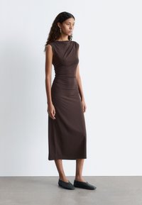 Braunes, figurbetontes, ärmelloses Kleid mit drapiertem Ausschnitt und Raffung in der Taille, kombiniert mit schwarzen flachen Schuhen auf einem glatten Boden.