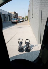 Paire de sandales noires à talons hauts avec brides à la cheville posées sur un trottoir vide à côté d’un bâtiment en briques grises dans une zone urbaine.