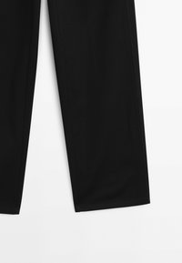Partie inférieure d’un pantalon noir à jambes droites sur fond blanc, montrant des ourlets propres et la texture du tissu.
