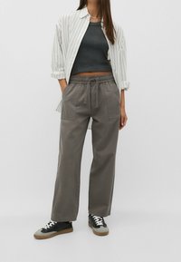 Nainen, joka on pukeutunut harmaisiin leveälahkeisiin housuihin, joissa on nauhavyötärö, mustaan lyhythihaiseen topiin, valkoisiin raidallisiin napillisiin paitaan ja musta-harmaisiin lenkkareihin.