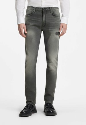 Mann trägt ausgewaschene schwarze Slim-Fit-Jeans mit einem kleinen abgenutzten Bereich am rechten Oberschenkel, kombiniert mit schwarzen Schuhen und einem weißen Hemd.