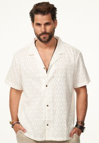 Kurze weiße Hemd mit geometrischem Muster, offenem Kragen, braunen Knöpfen und lässiger Passform, aus leichtem Stoff gefertigt.