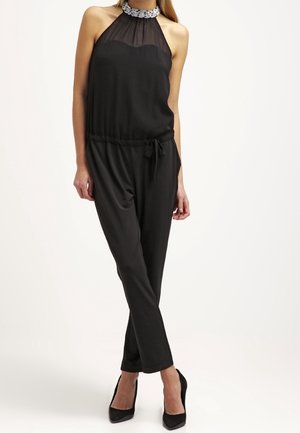 Frau trägt einen schwarzen Halterneck-Jumpsuit mit transparentem Yoke und verzierter Halskrause, kombiniert mit schwarzen spitzen High Heels, steht mit überschlagenen Beinen.
