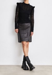 Femme portant un haut long à manches transparentes noir, une jupe en cuir noir arrivant aux genoux et des bottes à plateforme noires épaisses, se tenant contre un mur blanc.