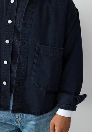 Torse d'un homme portant une veste sombre boutonnée avec une grande poche avant, superposée par-dessus un pull en tricot et un jean bleu clair.