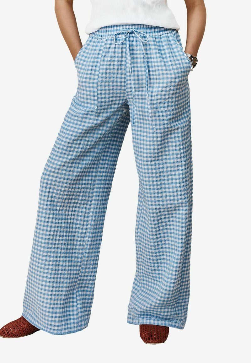 Personne portant un pantalon ample à carreaux bleu clair et blanc avec poches avant et taille à cordon, associé à des chaussures tressées marron.