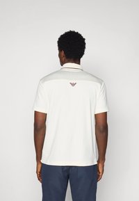 Κρέμα πόλο μπλούζα με κοντά μανίκια, γιακά και διακριτό υφαντό μοτίβο. Διαθέτει ένα μικρό καφέ λογότυπο στη πάνω πλάτη.