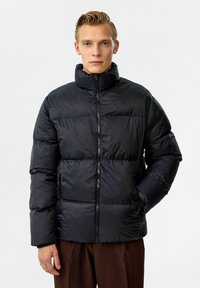Veste rembourrée noire avec un col haut, dotée d'une fermeture éclair à l'avant, de coutures horizontales et d'une texture lisse et brillante. Agrémentée de poches latérales.