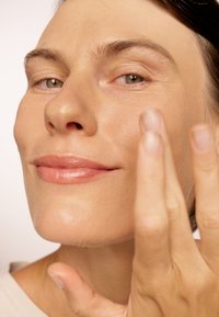 Nærbilde av en person som påfører hudpleie. Viser jevn, hydrert hud, nøytral leppefarge og middels langt brunt hår. Naturlig belysning.