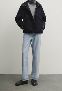 Manteau marine avec un col montant et une fermeture éclair, associé à un jean droit bleu clair et des chaussures noires. Tissu lisse, design minimaliste.