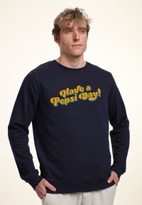 Sudadera azul marino de manga larga, con cuello redondo, y un gran gráfico amarillo y turquesa que dice "¡Ten un día de Pepsi!" en la parte frontal.