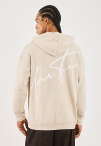 Mies, jolla on letitetyt hiukset, käyttää vaaleaa hupparia, jossa on valkoinen "Your Turn" -teksti takana, seisten vaaleaa taustaa vasten.