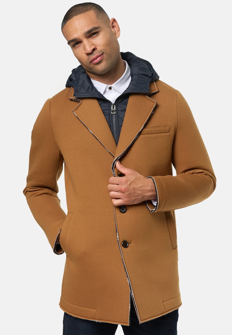 Brun kappa med en texturerad yta, som har ett navyfärgat, huven inre lager, utskurna slag och två framfickor. Knäppning med knappar och kontrasterande sömmar.