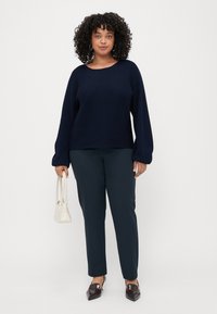Pull en maille surdimensionné bleu marine avec texture côtelée, associé à un pantalon slim-fit vert foncé. Le mannequin tient un petit sac à main crème et porte des chaussures à bout pointu.