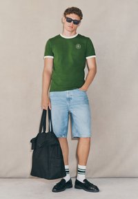Maglietta verde con bordi bianchi, pantaloncini di jeans, scarpe nere e borsa nera. Accessori comprendono occhiali da sole neri e calzini a righe.