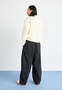 Κρέμα φούτερ με κατεβασμένα ώμου, σε συνδυασμό με χαλαρές μαύρες παντελόνες με δύο πίσω τσέπες, απεικονιζόμενο από πίσω. Απαλή φόντο.