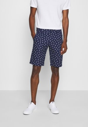 Marineblå shorts lavet af stof, med et hvidt geometrisk mønster, flad forside, sidelommer og en afslappet pasform. Bæret med en hvid t-shirt.