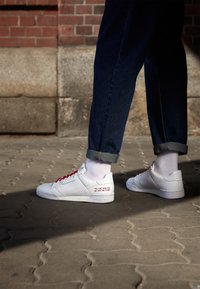 Sneaker bianchi con lacci rossi, in pelle, con lati perforati e un messaggio stampato sul tallone, abbinati a jeans scuri arrotolati alla caviglia.